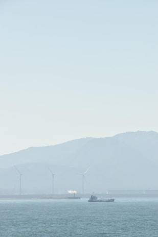 A serene seascape with a ship sailing on calm waters. In the background, several wind turbines stand against a hazy mountainous backdrop, and a small white plume of smoke or steam rises from a structure.
