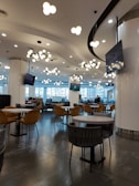 A modern cafe or lounge area with multiple round tables and plush chairs. The ceiling features numerous round, clustered light fixtures, creating an elegant atmosphere. The space is open and well-lit with large windows and a few people seated in the background, suggesting a quiet and relaxed environment.