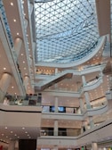 A modern shopping mall interior with multiple levels and a glass domed ceiling. The architecture includes numerous balconies and escalators, with a symmetrical arrangement of lights and pillars. People are visible on different floors, engaging in various activities, contributing to a lively atmosphere.