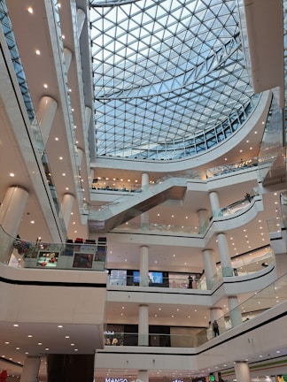 A modern shopping mall interior with multiple levels and a glass domed ceiling. The architecture includes numerous balconies and escalators, with a symmetrical arrangement of lights and pillars. People are visible on different floors, engaging in various activities, contributing to a lively atmosphere.