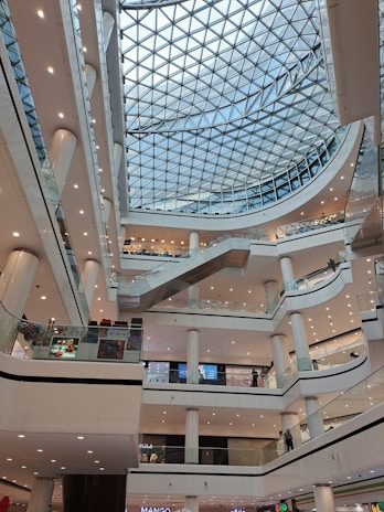 A modern shopping mall interior with multiple levels and a glass domed ceiling. The architecture includes numerous balconies and escalators, with a symmetrical arrangement of lights and pillars. People are visible on different floors, engaging in various activities, contributing to a lively atmosphere.