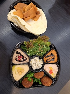 A platter with a variety of Middle Eastern dishes arranged in separate sections. The platter includes dishes like hummus with chickpeas and olive oil, tabbouleh salad with fresh parsley, falafel, and several sauces or dips garnished with ingredients like pomegranate seeds. Accompanying the platter is a plate of pita bread and toasted pita chips.