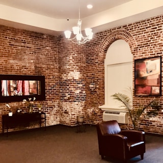 A cozy interior with exposed brick walls features a modern chandelier hanging from the ceiling. A leather armchair is placed on a carpeted floor next to a potted plant. A framed abstract painting and a decorative mirror add artistic flair to the room. A console table holds several decorative items, including vases and candles, and an air conditioning unit is built into one wall.