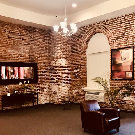 A cozy interior with exposed brick walls features a modern chandelier hanging from the ceiling. A leather armchair is placed on a carpeted floor next to a potted plant. A framed abstract painting and a decorative mirror add artistic flair to the room. A console table holds several decorative items, including vases and candles, and an air conditioning unit is built into one wall.