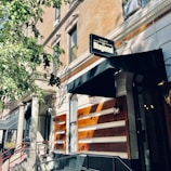 A street view of a building with a sign reading 'Agaveria La Diagonal' above a black awning. The building features traditional architecture with columns and large windows. A tree partially shades the facade, creating patterns of light and shadow on the building's beige and brown exterior.