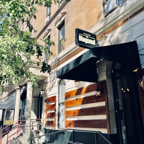A street view of a building with a sign reading 'Agaveria La Diagonal' above a black awning. The building features traditional architecture with columns and large windows. A tree partially shades the facade, creating patterns of light and shadow on the building's beige and brown exterior.
