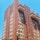 A tall red-brick apartment building with numerous windows, some featuring awnings. It has multiple protruding balconies and a green sign hanging over the side. The building is set against a clear blue sky.