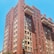 A tall red-brick apartment building with numerous windows, some featuring awnings. It has multiple protruding balconies and a green sign hanging over the side. The building is set against a clear blue sky.