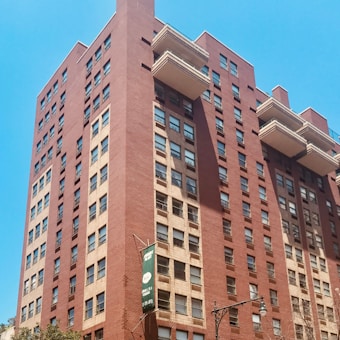 A tall red-brick apartment building with numerous windows, some featuring awnings. It has multiple protruding balconies and a green sign hanging over the side. The building is set against a clear blue sky.