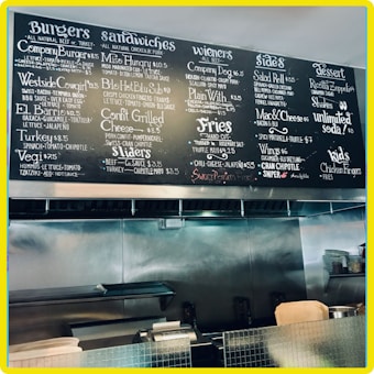 A restaurant menu board featuring various food options such as burgers, sandwiches, wieners, sides, desserts, and special items. The board has a chalkboard style design with handwritten items and prices, organized into clear categories. The background shows a kitchen area with stainless steel surfaces and cooking equipment.