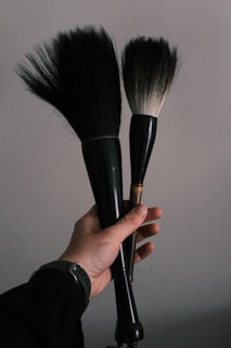 A hand holding two large, black-handled brushes against a plain background. The brushes have densely packed bristles, one slightly smaller than the other, and appear to be makeup or art brushes.