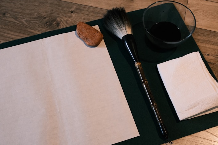 A traditional calligraphy setup is arranged on a wooden surface, featuring a sheet of blank paper, a large calligraphy brush, a small dish of black ink, a stack of paper towels, and an ink stone. The items suggest preparation for a writing or painting activity.