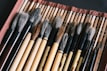 A collection of traditional calligraphy brushes with wooden handles laid out on a woven mat. The brushes vary in size and color, with soft bristles that range from white to dark brown. The mat has a red border, adding a contrasting color to the natural tones of the brushes.