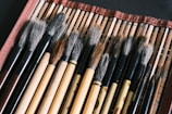 A collection of traditional calligraphy brushes with wooden handles laid out on a woven mat. The brushes vary in size and color, with soft bristles that range from white to dark brown. The mat has a red border, adding a contrasting color to the natural tones of the brushes.