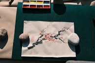 A piece of paper with an artistic illustration depicting a branch with red blossoms and dark ink strokes, held down by two stones on a green mat. A watercolor palette with various colors is positioned above the paper.