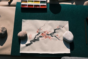 A piece of paper with an artistic illustration depicting a branch with red blossoms and dark ink strokes, held down by two stones on a green mat. A watercolor palette with various colors is positioned above the paper.