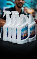 Several white bottles of hookah cleaner are arranged in a row on a dark surface. Each bottle has a spray nozzle and a blue label with the product name in bold letters. A blurred figure of a person appears in the background, partially visible and out of focus. The setting suggests an indoor environment, likely a store or exhibition.