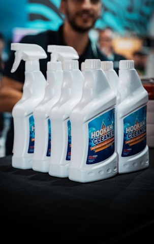 Several white bottles of hookah cleaner are arranged in a row on a dark surface. Each bottle has a spray nozzle and a blue label with the product name in bold letters. A blurred figure of a person appears in the background, partially visible and out of focus. The setting suggests an indoor environment, likely a store or exhibition.