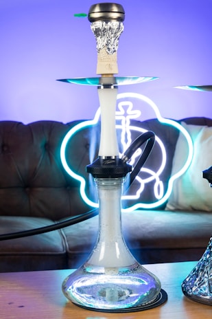 A hookah with a glass base is placed on a wooden table. The background features a brown sofa with a neon sign that emits a blue and white glow. The smoke can be seen coming from the top of the hookah, which is covered with foil.