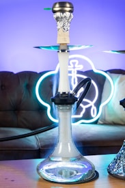 A hookah with a glass base is placed on a wooden table. The background features a brown sofa with a neon sign that emits a blue and white glow. The smoke can be seen coming from the top of the hookah, which is covered with foil.