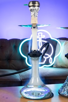 A hookah with a glass base is placed on a wooden table. The background features a brown sofa with a neon sign that emits a blue and white glow. The smoke can be seen coming from the top of the hookah, which is covered with foil.