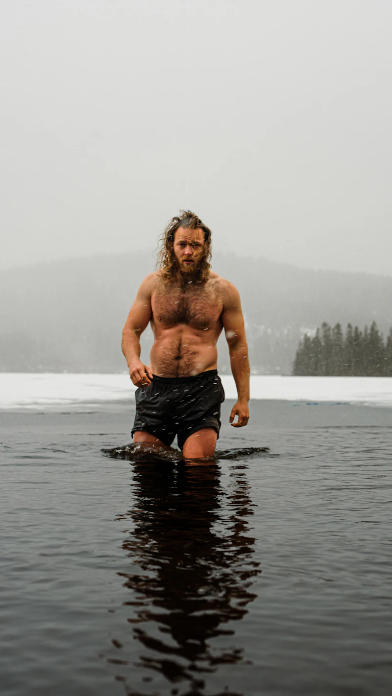 Homem sem camisa, de shorts e com cabelo comprido e barba molhados, em pé até a cintura em um lago congelado, com neve e pinheiros ao redor