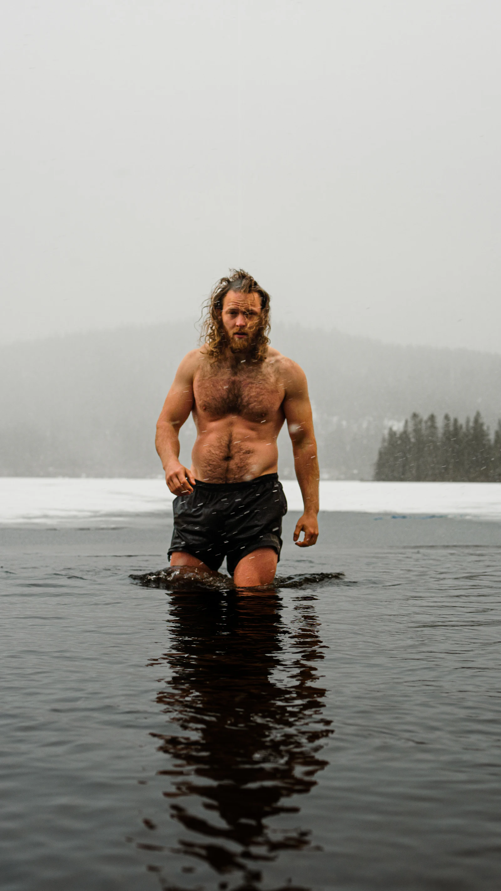 Homem sem camisa, de shorts e com cabelo comprido e barba molhados, em pé até a cintura em um lago congelado, com neve e pinheiros ao redor