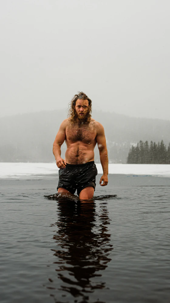Homem sem camisa, de shorts e com cabelo comprido e barba molhados, em pé até a cintura em um lago congelado, com neve e pinheiros ao redor