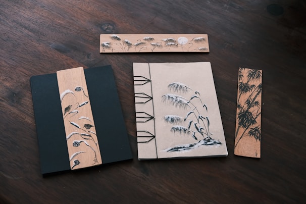 A collection of artistic wooden bookmarks and a notebook placed on a dark wooden surface. The bookmarks are decorated with intricate ink drawings featuring scenes of birds, bamboo, and reeds. The notebook features a similar style of artwork with binding details visible on the side.