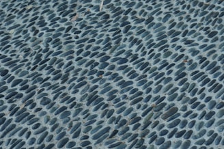 A close-up view of a textured surface featuring numerous smooth, oval stones embedded in concrete. The stones are dark grey and arranged in a pattern that creates a sense of movement and rhythm across the surface.