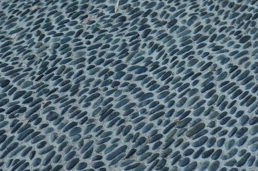 A close-up view of a textured surface featuring numerous smooth, oval stones embedded in concrete. The stones are dark grey and arranged in a pattern that creates a sense of movement and rhythm across the surface.