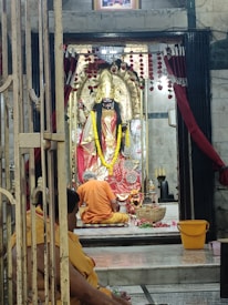A religious scene within a temple featuring a decorated idol dressed in ornate garments and jewelry. The idol is adorned with garlands and sits inside a shrine that is framed by red and gold decorations. In front of the idol is a person seated, possibly engaged in prayer or offering rituals. The area is lit softly, enhancing the sacred atmosphere.