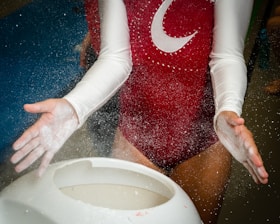 A person clad in a red leotard with white sleeves applies chalk to their hands above a white chalk bowl. Chalk particles are visibly dispersing into the air, demonstrating preparation and focus.
