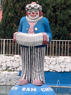 A large colorful clown statue with a cheerful face, wearing a blue jacket, striped pants, and holding an accordion. It is set against a background of lush green trees and a white stone wall with a blue-painted base area at the bottom.
