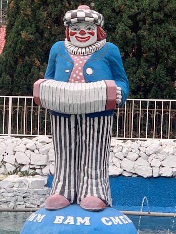 A large colorful clown statue with a cheerful face, wearing a blue jacket, striped pants, and holding an accordion. It is set against a background of lush green trees and a white stone wall with a blue-painted base area at the bottom.