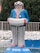 A large colorful clown statue with a cheerful face, wearing a blue jacket, striped pants, and holding an accordion. It is set against a background of lush green trees and a white stone wall with a blue-painted base area at the bottom.