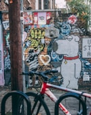 A vibrant urban street art scene with a mix of colorful graffiti and murals on a brick wall. The artwork includes abstract designs, cartoon characters like a mosaic dog, and various stickers and posters. A red and white bicycle is parked in the foreground, suggesting an urban environment.