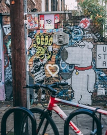 A vibrant urban street art scene with a mix of colorful graffiti and murals on a brick wall. The artwork includes abstract designs, cartoon characters like a mosaic dog, and various stickers and posters. A red and white bicycle is parked in the foreground, suggesting an urban environment.