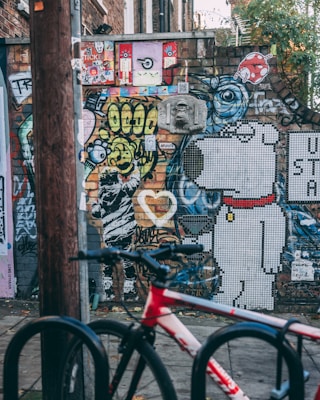 A vibrant urban street art scene with a mix of colorful graffiti and murals on a brick wall. The artwork includes abstract designs, cartoon characters like a mosaic dog, and various stickers and posters. A red and white bicycle is parked in the foreground, suggesting an urban environment.