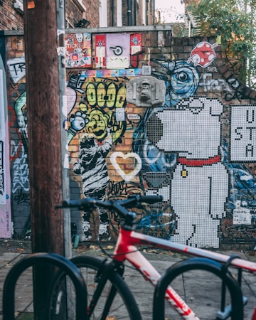 A vibrant urban street art scene with a mix of colorful graffiti and murals on a brick wall. The artwork includes abstract designs, cartoon characters like a mosaic dog, and various stickers and posters. A red and white bicycle is parked in the foreground, suggesting an urban environment.