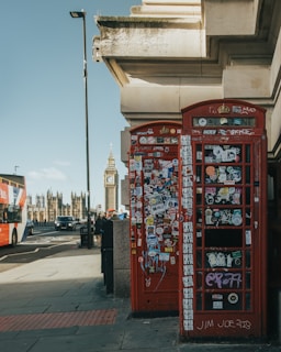 A red telephone booth covered in colorful stickers stands on a city street. In the background, the iconic clock tower of Big Ben and the Houses of Parliament are visible. A double-decker bus and several cars are seen on the road nearby, with pedestrians walking along the sidewalk. The scene captures a mix of urban life and historical architecture under a clear blue sky.