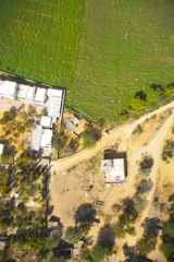 Aerial view of a rural landscape with a mix of agricultural fields and residential structures. The image features a large green field on one side, bordered by buildings, pathways, and scattered vegetation and trees. Dirt roads connect the various structures and sections of the area.