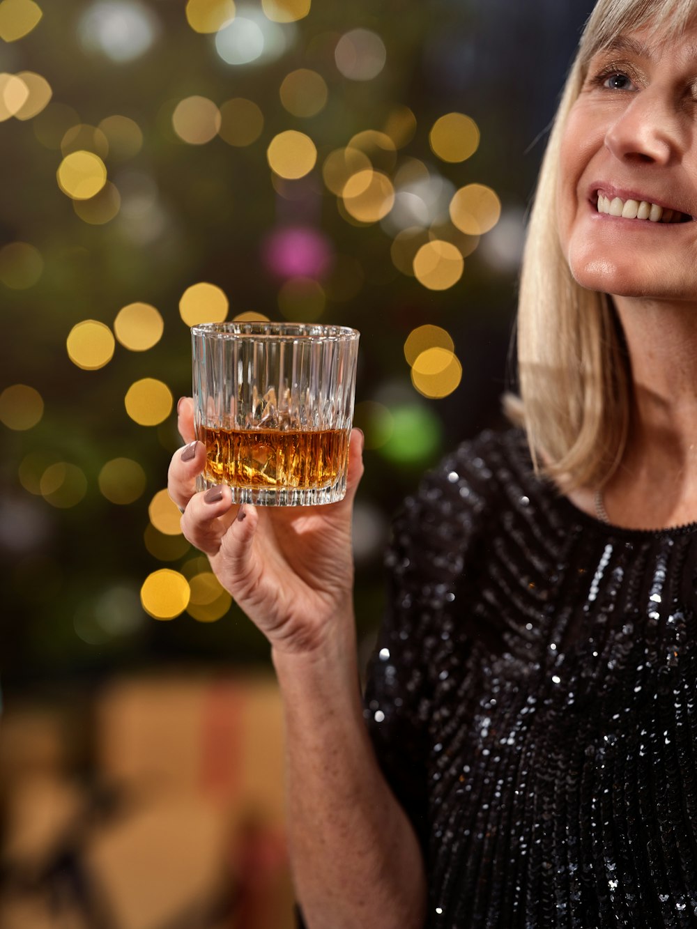 a woman holding a glass of whiskey in front of a christmas tree