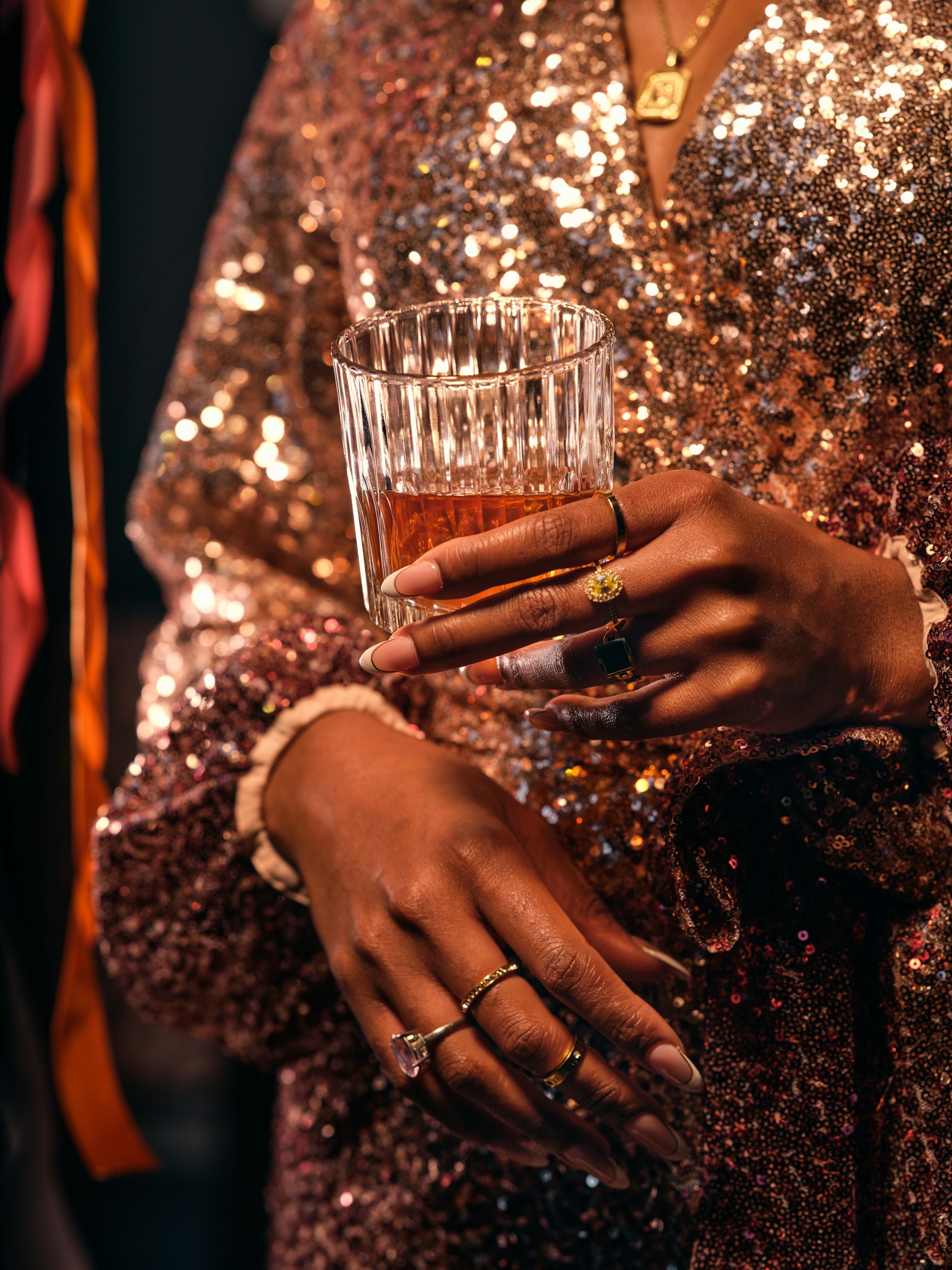 A close-up of a person wearing a shimmering, sequined dress, holding a glass filled with a golden-brown liquid. The person's hands are adorned with multiple rings, showcasing elegant jewelry. Warm lighting highlights the sparkles in the fabric, creating a festive, luxurious vibe.