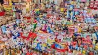 A vast array of packaged food products displayed on shelves, showcasing a variety of snacks, beverages, canned goods, and pantry staples. The arrangement includes brands from different categories such as candy, crackers, cereal, sauces, and bottled drinks. The scene is colorful and packed tightly, illustrating a well-stocked grocery or convenience store.