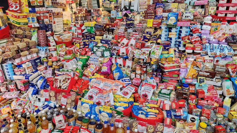A vast array of packaged food products displayed on shelves, showcasing a variety of snacks, beverages, canned goods, and pantry staples. The arrangement includes brands from different categories such as candy, crackers, cereal, sauces, and bottled drinks. The scene is colorful and packed tightly, illustrating a well-stocked grocery or convenience store.