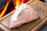 A raw piece of meat topped with a sprig of rosemary lies on a wooden cutting board. In the background, there is a bright flame, suggesting a fireplace or a grilling station. Another sprig of herbs is placed to the side on the cutting board.