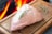 A raw piece of meat topped with a sprig of rosemary lies on a wooden cutting board. In the background, there is a bright flame, suggesting a fireplace or a grilling station. Another sprig of herbs is placed to the side on the cutting board.