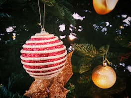 A Christmas tree adorned with various ornaments, including a large red and white striped bauble, a smaller golden bauble, and a decorative golden ribbon. The green branches of the tree and surrounding lights create a festive atmosphere.