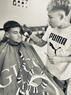 A person with tattoos and a Puma T-shirt is styling the hair of another individual who is wearing a barber cape with a snake design and the Gucci logo. They appear focused on a precision haircut. The room is equipped with chairs and a poster on the wall showing different hairstyles.